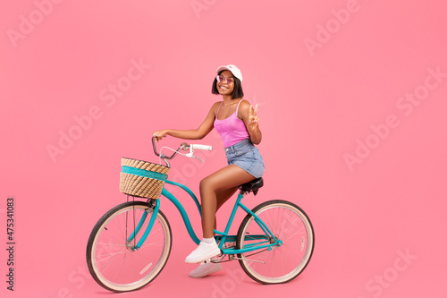 Summer activity. Young black lady in cool outfit riding bicycle, showing peace gesture on pink background, full length © Prostock-studio