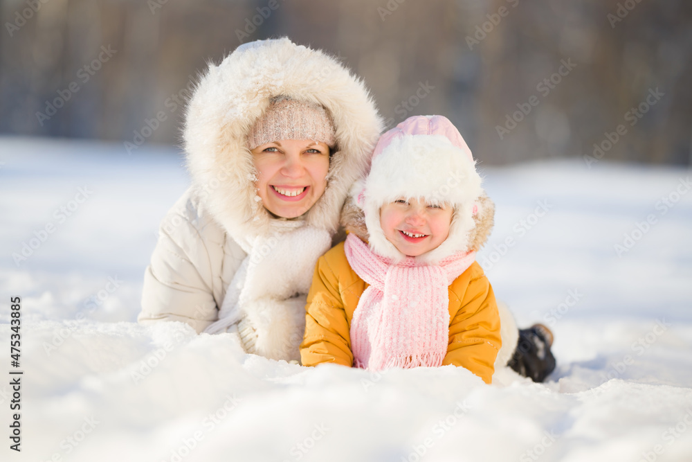 Happy young mother and little daughter smiling and lying down on white snow at nature park. Lovely emotional moment. Spending time together in beautiful cold sunny winter day. Front view. Closeup. Happy young mother and little daughter smiling and lying down on white snow at nature park. Lovely emotional moment. Spending time together in beautiful cold sunny winter day. Front view. Closeup.