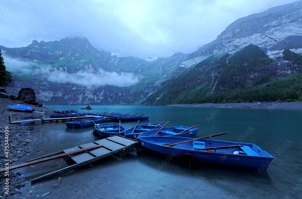 Morning scenery of peaceful Lake Oeschinensee before dawn with rowboats parking by the lakeside & foggy mountains in background under moody cloudy sky in blue twilight in Berner Oberland, Switzerland