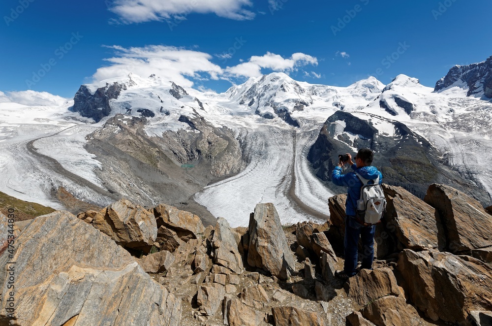 Tourist standing at the edge of a rocky cliff, taking photos of impressive Gorner Glacier & majestic snow capped mountains in background under blue sunny sky in Gornergrat, Zermatt, Valais Switzerland