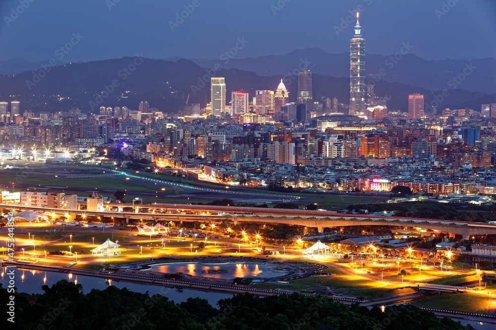 Aerial panorama of Taipei in evening twilight, capital city of Taiwan, with Taipei 101 Tower amid skyscrapers in downtown, a riverside park by Keelung River & an airplane parking in Songshan Airport