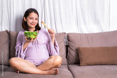 Happy woman holding glass bowl with fresh salad. Eating healthy vegetable organic food . Care about life. sitting on sofa in living room. Copy space for text.