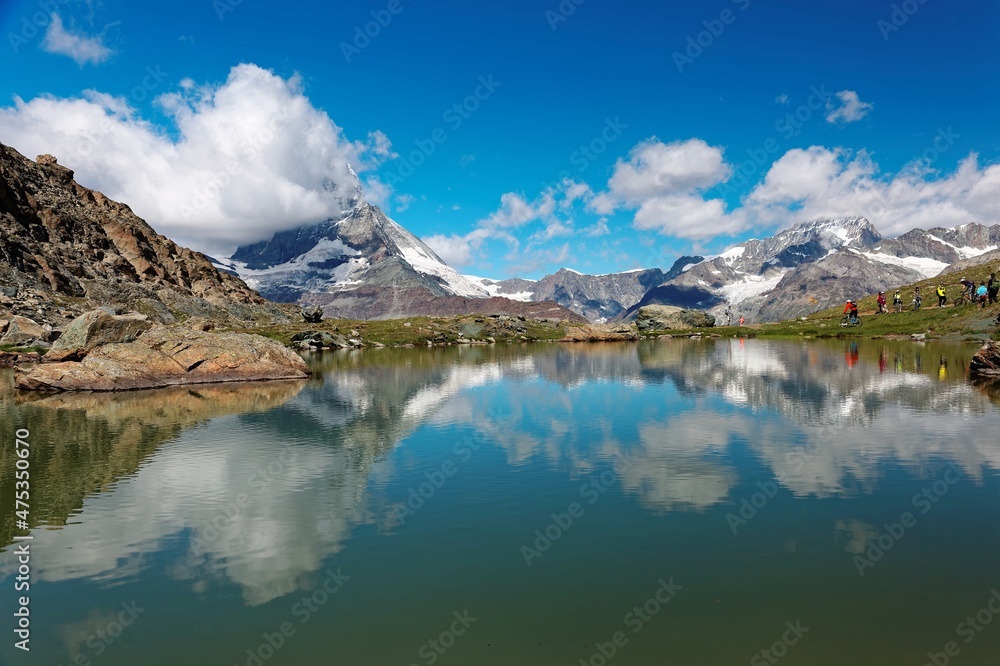 Tourists hiking and mountain biking by Lake Riffelsee on a beautiful sunny summer day with Mountain Matterhorn under blue sky reflected on the peaceful water in Zermatt, Valais, Switzerland, Europe