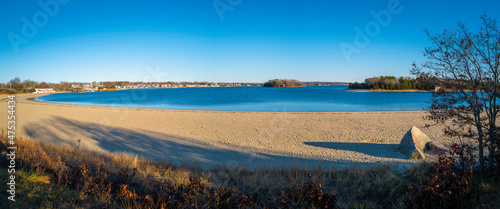 Panoramic curved shorelines with the views of blue sea and sky, trees, rock, and coastal village. Tranquil seascape over the Onset Bay and Beach in Onset, Massachusetts in winter.