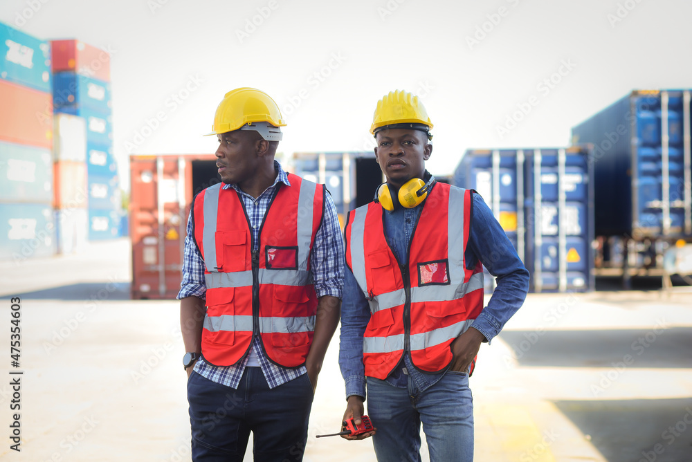 Two African black male worker standing and wearing safety work equipment clothes is Working and checking containers at a warehouse, ship, cargo, import, export  industry.