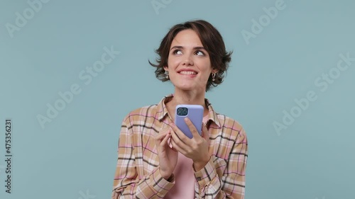 Dreamful pensive minded young brunette woman 20s years old wears plaid shirt hold using mobile cell phone typing browsing chat send sms isolated on pastel plain light blue background studio portrait