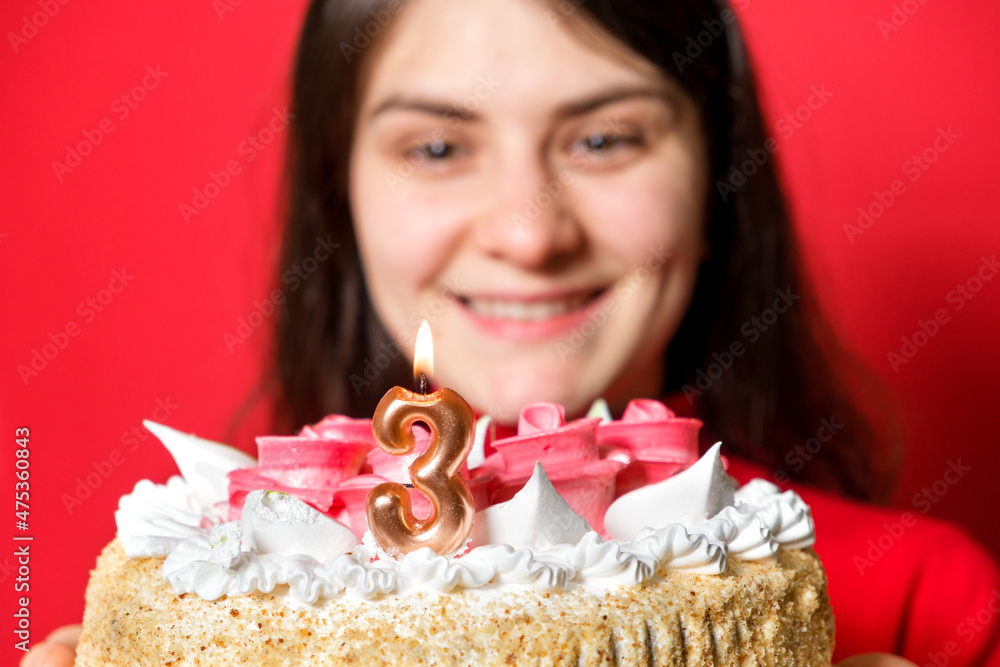 A happy woman holds a cake with a candle in the shape of the number three on a red background. A happy woman holds a cake with a candle in the shape of the number three on a red background.