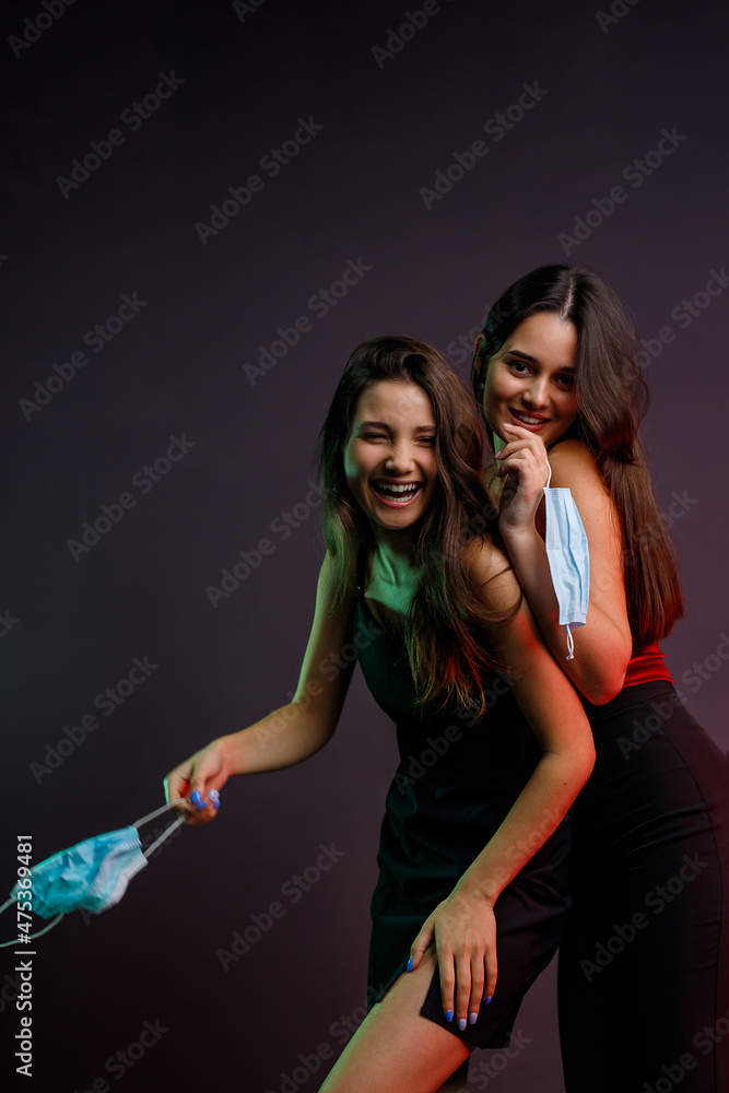 Two exited young women in evening dresses and manicure happy having fun and keeping in their hands medical masks to go to the party on pandemic time. Two exited young women in evening dresses and manicure happy having fun and keeping in their hands medical masks to go to the party on pandemic time.