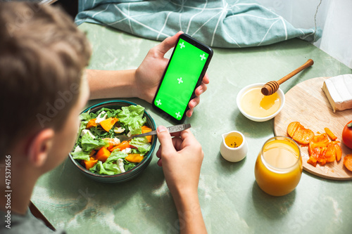 Concept of calorie counting, dietary nutrition online shopping and online training. A teenage boy has breakfast with a healthy vitamin salad and holds a phone with a green chroma key screen.