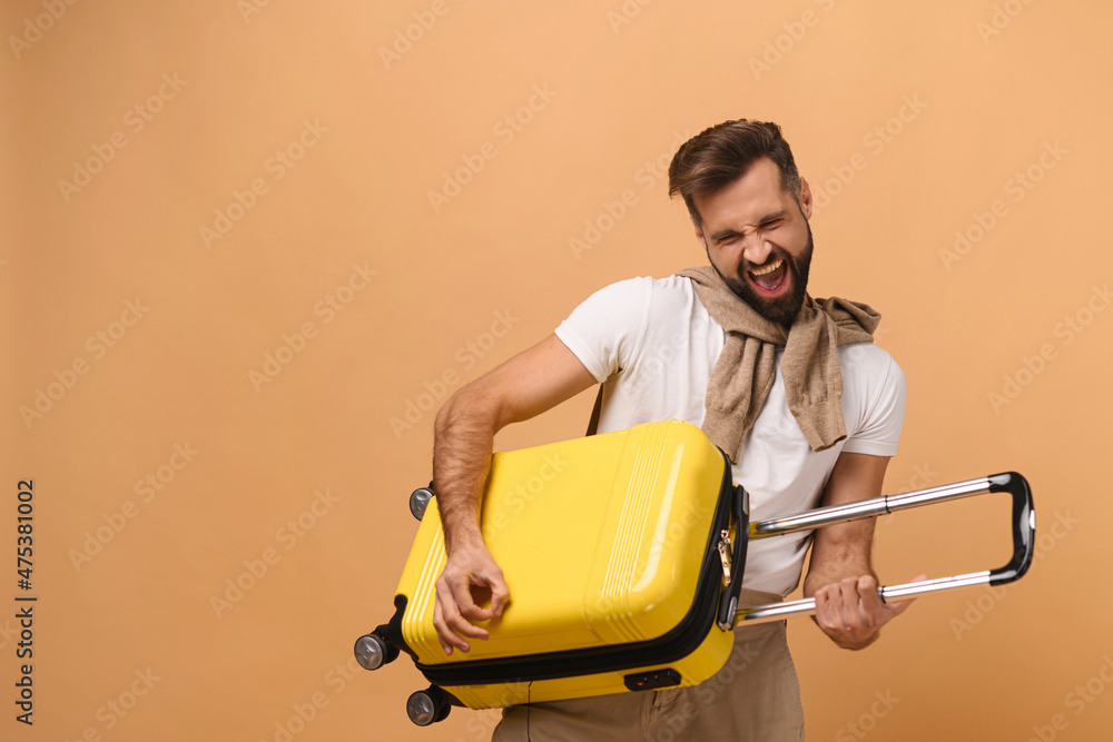 Cheerful caucasian man standing with yellow suitcase, playing on a bag like a guitarist, fooling around, while looking forward a trip isolated on orange background Cheerful caucasian man standing with yellow suitcase, playing on a bag like a guitarist, fooling around, while looking forward a trip isolated on orange background