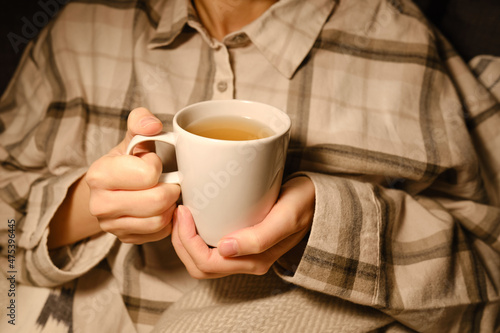 A cup of tea in the hands of a girl. The girl is drinking hot tea. A girl in pajamas is wrapped in a blanket and is drinking delicious tea. Enjoy the comforts of home. White cup close up in woman hand