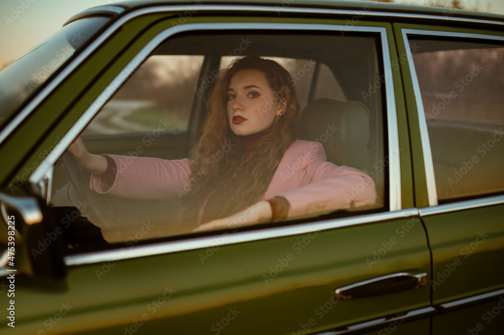 Fashionable confident woman posing at the wheel of green retro car. Elegant redhead model wearing classic pink suit. Copy, empty space for text Fashionable confident woman posing at the wheel of green retro car. Elegant redhead model wearing classic pink suit. Copy, empty space for text
