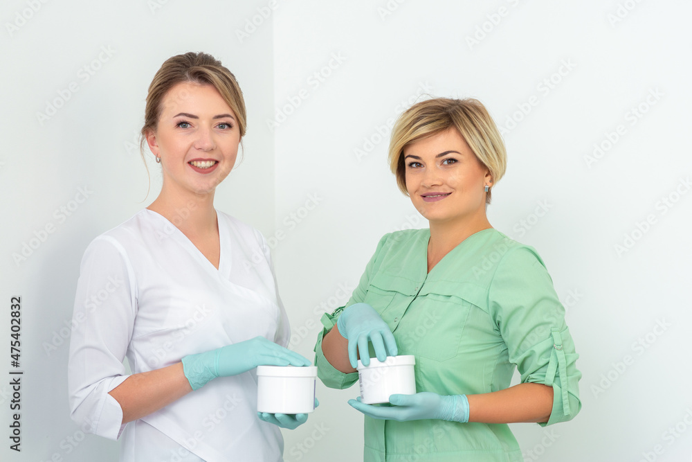 Two cosmeticians with jars of wax for depilation smiling against a white background. Natural product for hair removal. Copy space Two cosmeticians with jars of wax for depilation smiling against a white background. Natural product for hair removal. Copy space