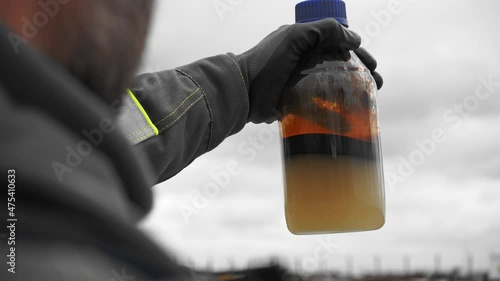 An OIL WORKER holds up a SAMPLE of CRUDE OIL in a plastic bottle for field inspection. Outdoor close-up of petroleum extraction analysis.