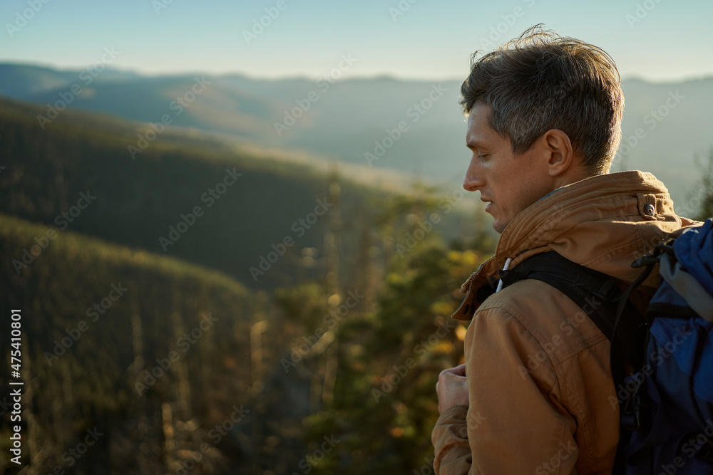 Side close up portrait man traveler with backpack standing on mountain top at sunset, hiker looking confident and happy with his hiking expedition outdoors.