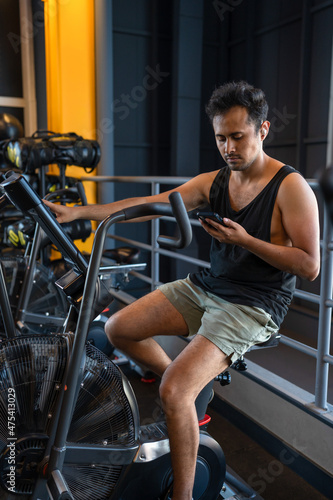 Young man cheking his phone while he is on a cycling machine in the gym