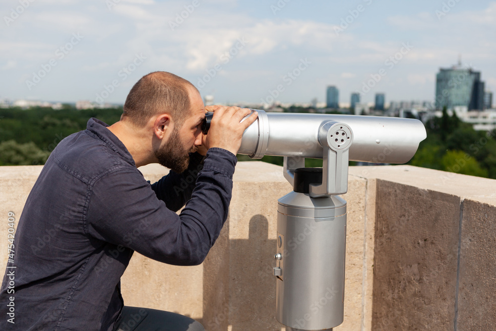 Caucasian male wathching through binoculars telescope standing on tower rooftop at observation point. Tourist enjoying metropolitan urban city landscape. Travel concept. Panoramic view