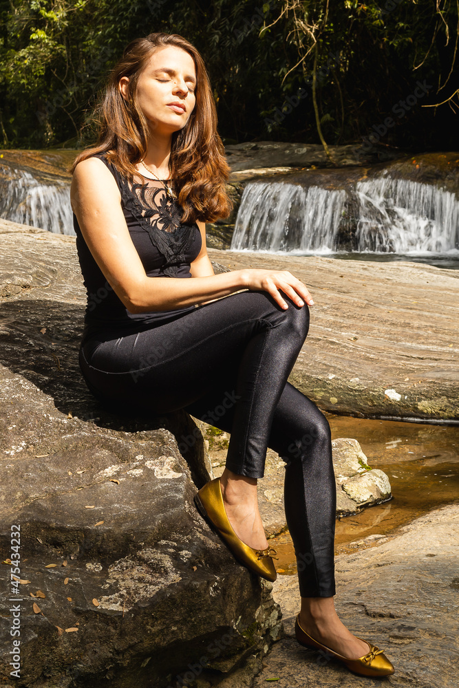 Jovem mulher de pele clara com cabelos loiros e roupas pretas sentada nas rochas à beira de uma pequena cachoeira de águas límpidas com o sol no rosto Jovem mulher de pele clara com cabelos loiros e roupas pretas sentada nas rochas à beira de uma pequena cachoeira de águas límpidas com o sol no rosto