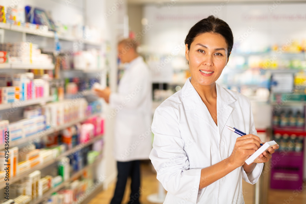 Asian female pharmacist in lab coat standing in salesroom of drugstore and writing recipe in blank.