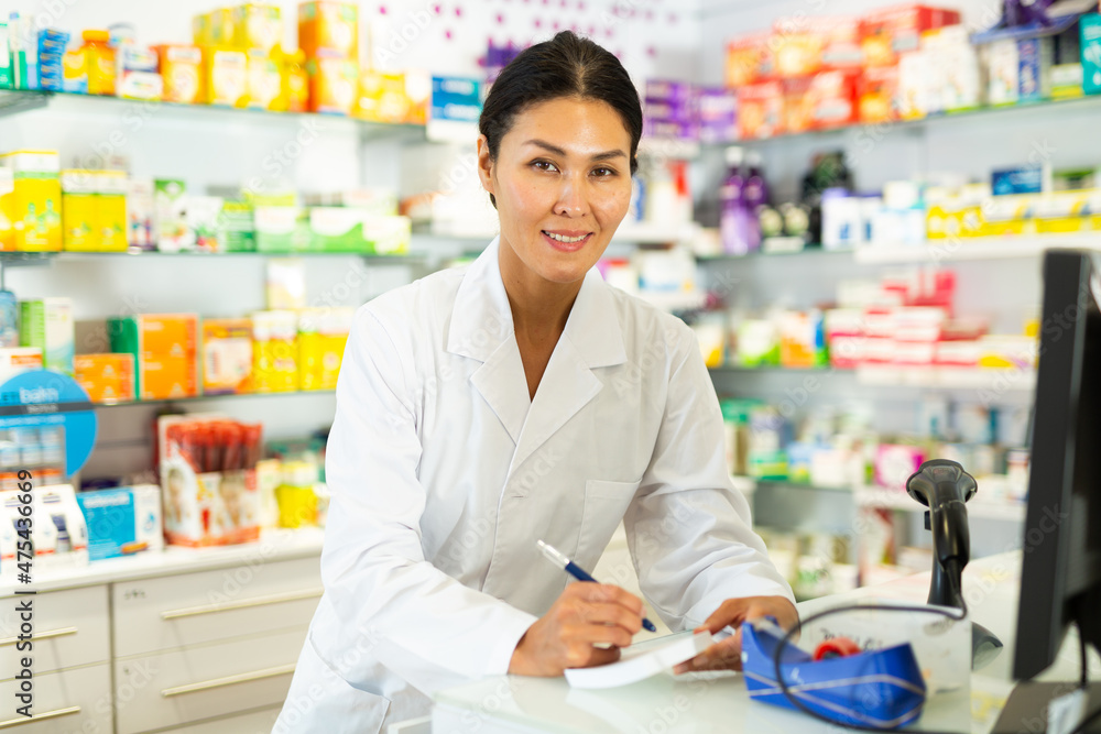 Asian female pharmacist in lab coat standing beside counter in drugstore and writing.