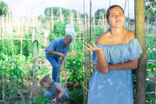 Family of three in kitchen garden. Mother looks unhappy and annoyed and standing in foreground. Father and daughter working in background.