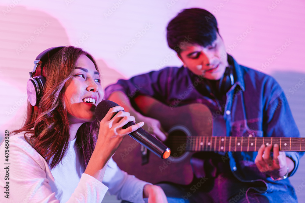 Obraz premium Pretty Asian female singer recording songs by using a studio microphone with male playing guitar in blue and red light. Performance and show in the music business. Duet session.