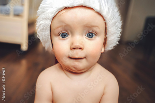 Bild auf Leinwand A small big-eyed surprised child, newborn, baby or toddler in a Santa Claus hat is sitting on the floor at home