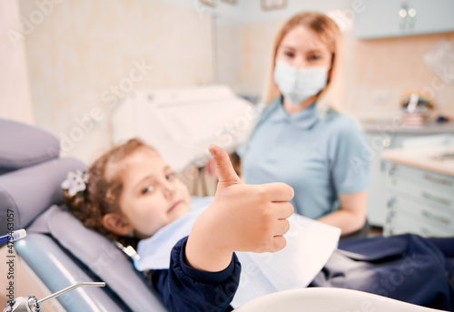 Focus on girl s hand giving thumbs up while woman stomatologist in medical mask sitting beside kid in dental chair. Concept of pediatric dentistry and dental care approval.