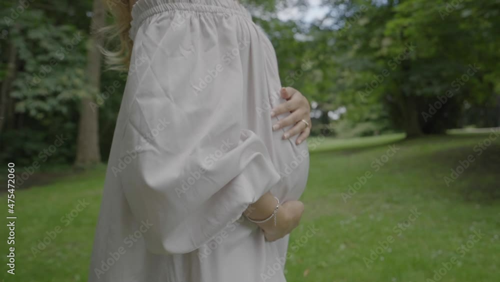 Closeup shot of a pregnant young woman wearing a beautiful white dress and holding her large belly, while walking through her lush green garden Closeup shot of a pregnant young woman wearing a beautiful white dress and holding her large belly, while walking through her lush green garden