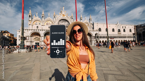 Audio tour online app on digital mobile smartphone. Happy young student woman holding phone listening audioguide at San Marco square in Venice, Italy. Simultaneous translation devices.