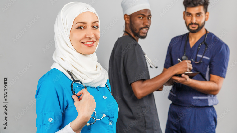 Portrait of positive healthcare workers standing in the hallway and looking at the camera. Group of medics smiling over gray background. muslim doctor woman on background of african and indian doctor