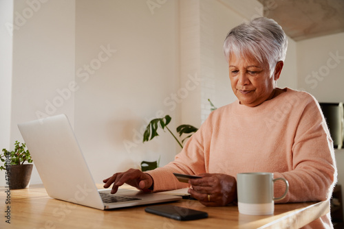 Elderly multi-ethnic female happily typing in bank details to make online payment on laptop, sitting at kitchen counter. 