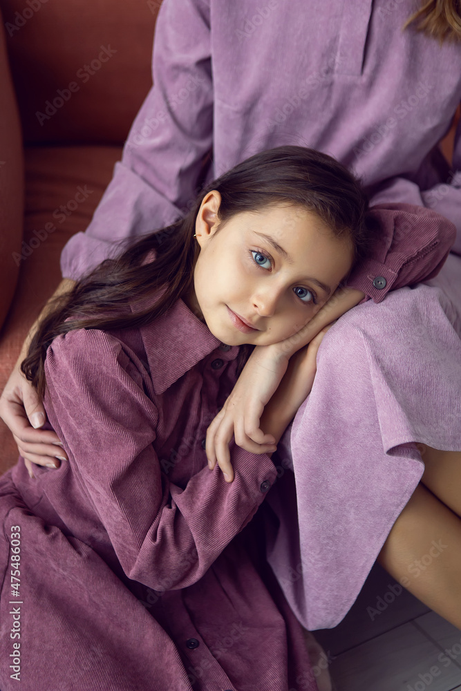 Caucasian girl with big eyes in a pink dress is sitting on a sofa with a woman mother with a Christmas tree in a white studio room with light from the window Caucasian girl with big eyes in a pink dress is sitting on a sofa with a woman mother with a Christmas tree in a white studio room with light from the window