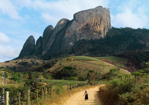 Fotografia turista caminhando em estrada com Pedra dos Cinco Pontões ao fundo, em Laranja d