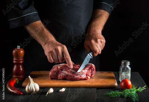 The chef cuts meat with knife in the kitchen prepares food. Vegetables and spices on the kitchen table in a restaurant to prepare a delicious lunch
