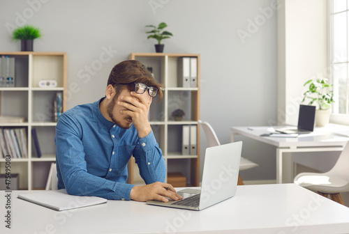 Stressful office job finally turns young man into a burnout. Stressed, tired, exhausted male worker overwhelmed with problems sitting at working desk with laptop computer and holding hand on his face