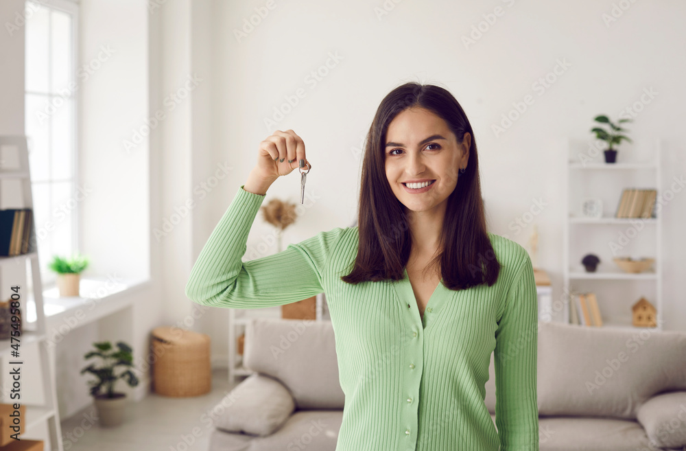 Portrait of beautiful young female homeowner showing keys to her new home. Caucasian brunette woman in green sweater posing in living room. Concept of real estate, moving home or renting apartment. Portrait of beautiful young female homeowner showing keys to her new home. Caucasian brunette woman in green sweater posing in living room. Concept of real estate, moving home or renting apartment.