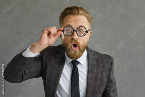 Portrait of shocked and funny caucasian businessman in glasses with magnifying glass. Man dressed in a stylish costume on a gray concrete background. Concept of considering new business ideas.