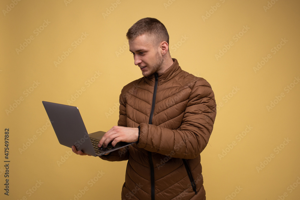Young 20s man in a brown jacket, on a yellow background, holding the use of work on a laptop, typing text, browsing in a chat, sending sms Young 20s man in a brown jacket, on a yellow background, holding the use of work on a laptop, typing text, browsing in a chat, sending sms