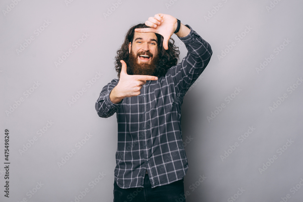 Positive bearded male in checkered shirt making frame gesture with hands and looking at camera on gray background in studio Positive bearded male in checkered shirt making frame gesture with hands and looking at camera on gray background in studio