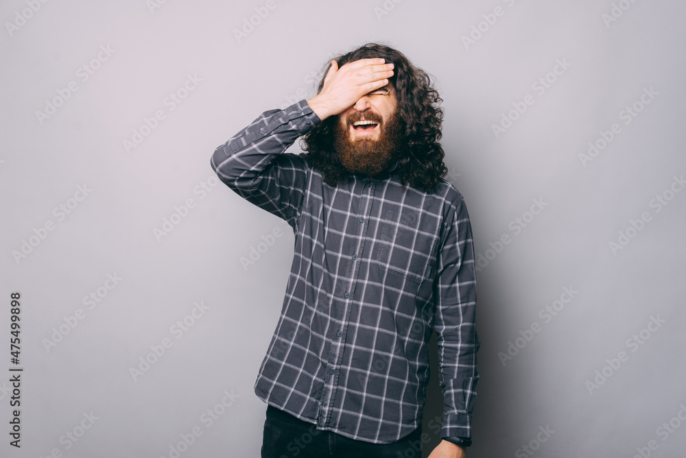 Confused bearded man in checkered shirt with closed eyes standing on gray background with hand on forehead in light studio Confused bearded man in checkered shirt with closed eyes standing on gray background with hand on forehead in light studio