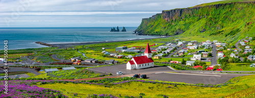 Billede på lærred Panoramic view of basalt stacks Reynisdrangar, volcanic black sand beach and violet lupine and yellow meadow flowers at Vik town, and a Lutheran church, South Iceland, at summer sunny day  blue sky