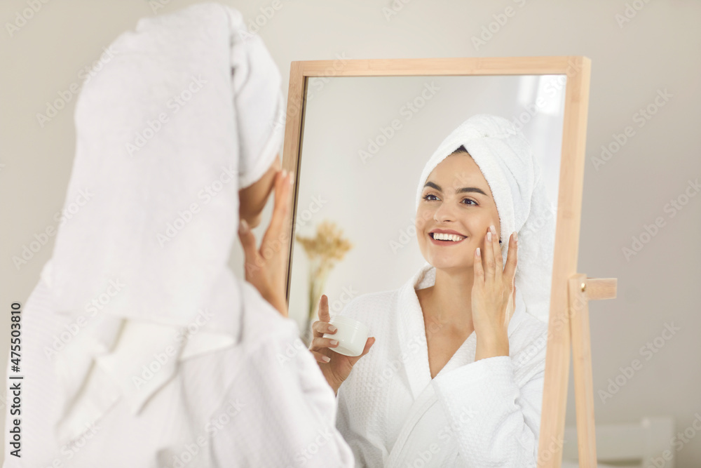 Portrait of smiling beautiful woman who after shower applies moisturizer on face. Lady smoothes cream on perfect skin in front of mirror in bathrobe and towel. Concept of daily morning beauty routine. Portrait of smiling beautiful woman who after shower applies moisturizer on face. Lady smoothes cream on perfect skin in front of mirror in bathrobe and towel. Concept of daily morning beauty routine.