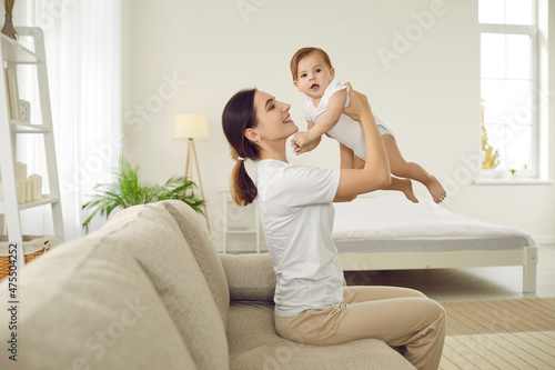 Portrait of happy cheerful young woman holding her baby and playing with her at home. Side view of woman sitting on sofa in bright bedroom and having fun lifting asix month old baby high in air.