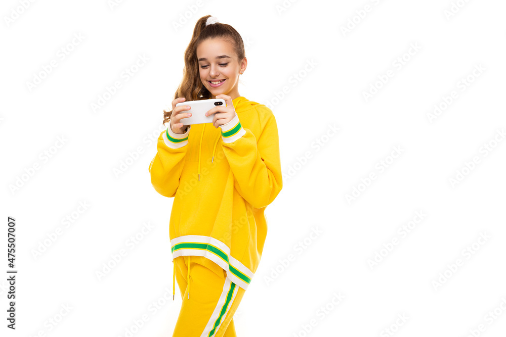 Photo of a beautiful smiling cheerful cute girl with dark hair with a ponytail in a stylish yellow tracksuit with green stripes plays a mobile phone with a white case isolated on a white background Photo of a beautiful smiling cheerful cute girl with dark hair with a ponytail in a stylish yellow tracksuit with green stripes plays a mobile phone with a white case isolated on a white background