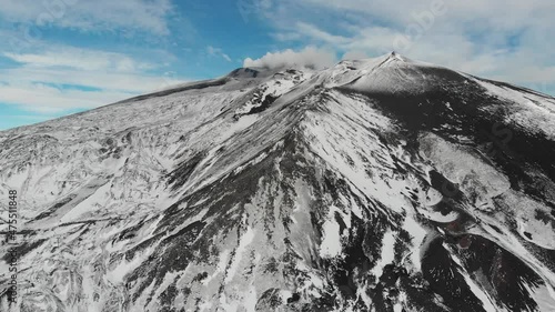 Drone on the side of mount etna descends on a snowy day, smoke can be seen erupting from main crater at the top