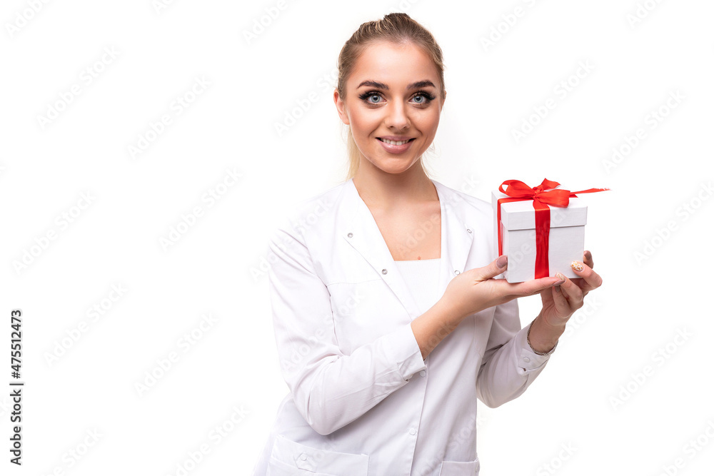 Photo of a beautiful pretty attractive positive smiling young blonde woman with a ponytail and makeup in a white medical coat of a nurse holding a white gift box with a red ribbon isolated on a white Photo of a beautiful pretty attractive positive smiling young blonde woman with a ponytail and makeup in a white medical coat of a nurse holding a white gift box with a red ribbon isolated on a white