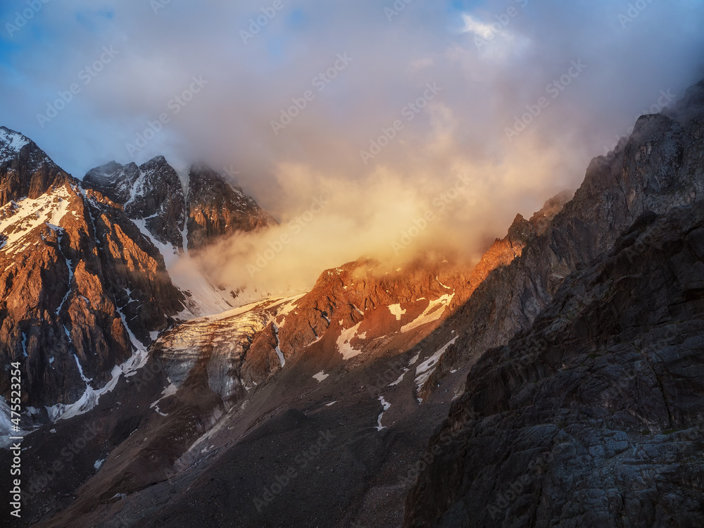 Sunlight in the mountains. Big glacier on top in orange light. Scenic mountain landscape with great snowy mountain range lit by dawn sun among low clouds.