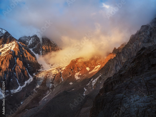 Sunlight in the mountains. Big glacier on top in orange light. Scenic mountain landscape with great snowy mountain range lit by dawn sun among low clouds.