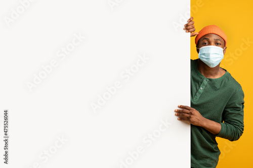 Black young man in medical mask standing behind blank white advertisement board